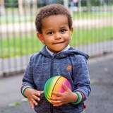 A young child stood in a playground wearing hearing aids and a grey cardigan with a hood is holding a colourful striped ball 