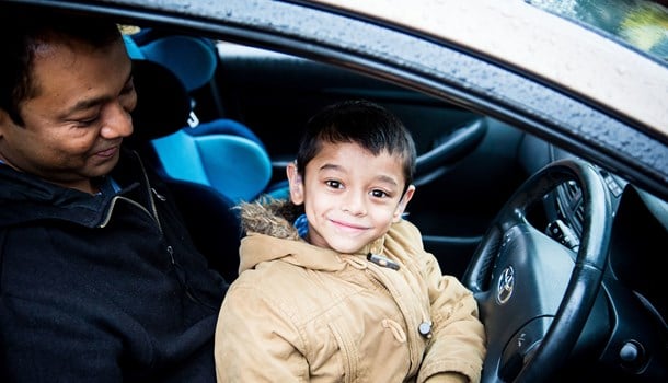 A boy wearing hearing aids smiling at the camera and sitting on his dad's lap in the car.