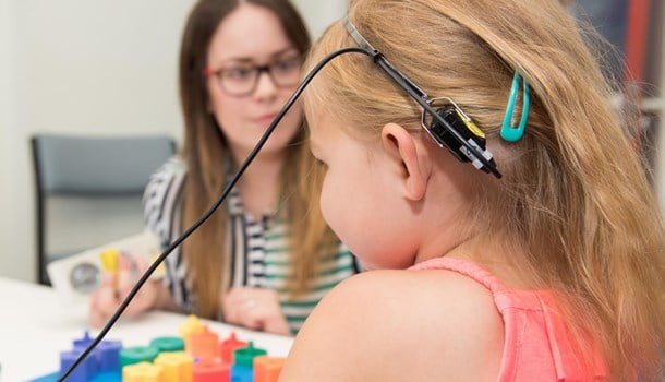 A young girl faces away wearing monitoring hearing equipment, and her audiologist is in the background.