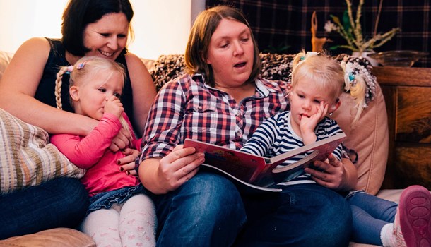 Mums sat on a sofa reading a picture book to their young twin daughters.