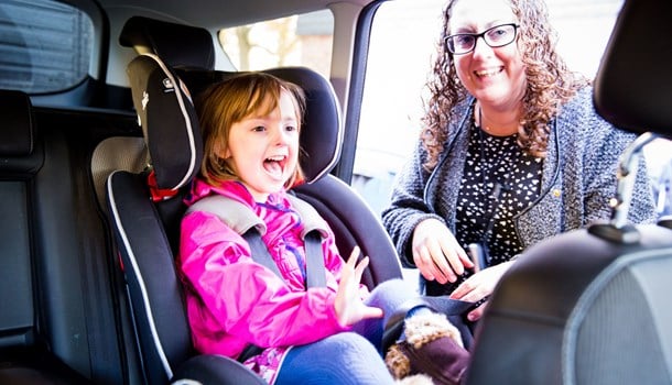 A deaf young girl smiling while her mum buckles her into a car seat.