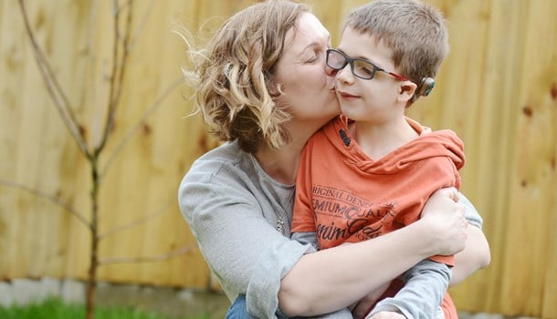 A boy with microtia and atresia wearing glasses and a bone-anchored hearing aid (BAHA) hugging his mum outside.