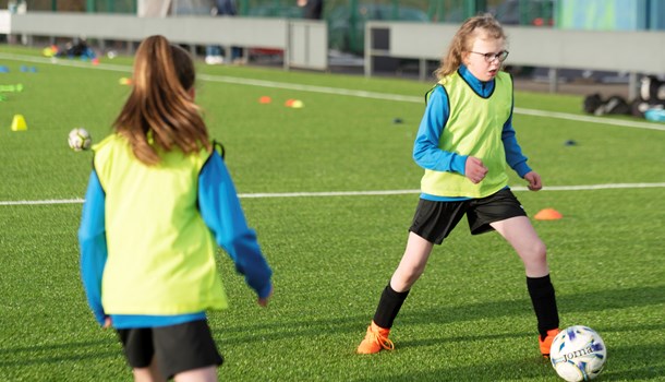 Daisy wearing a high-vis bib playing football with a girl on a football pitch.