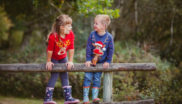 A deaf young boy wearing blue hearing aids and his sister stood on a fence wearing Christmas jumpers.