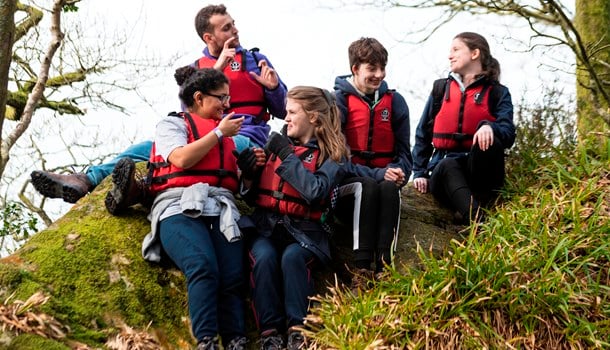 A group of deaf young people wearing life jackets sit on a grassy slope, chatting in sign language.