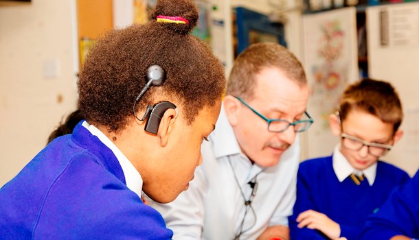 A teacher leans over to speak with two students, one of whom is wearing a cochlear implant.