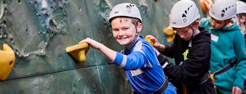 A boy wearing a blue top and a helmet is climbing along a climbing wall. He is smiling at the camera.